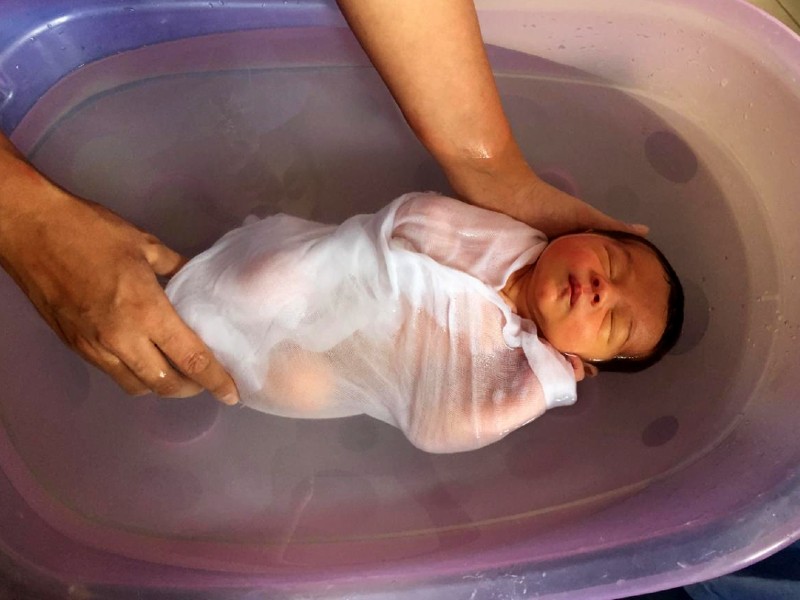 Bebê recém-nascido enrolado a uma fralda branca está deitado dentro de uma banheira, dormindo. Ele é apoiado por duas mãos que aparentam ser de uma mulher